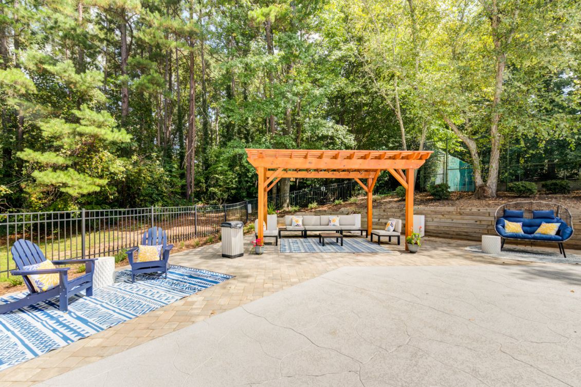 Outdoor lounge area with wooden pergola, stone seating, and blue cushioned chairs surrounded by mature trees at Madison Laurel Oaks apartments in North Raleigh, NC