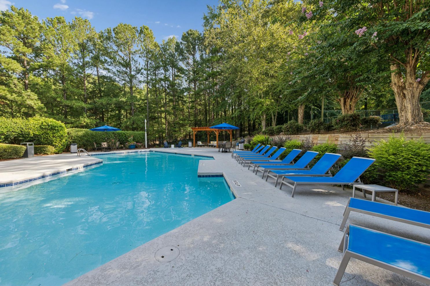 An outdoor swimming pool surrounded by blue lounge chairs, patio umbrellas, and trees.